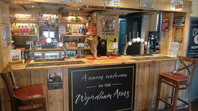 The bar at The Wyndham Arms, with hand pumps, bottles on the shelves and Loki the pub dog sitting behind the counter, above a chalkboard reading “A warm welcome to the Wyndham Arms”