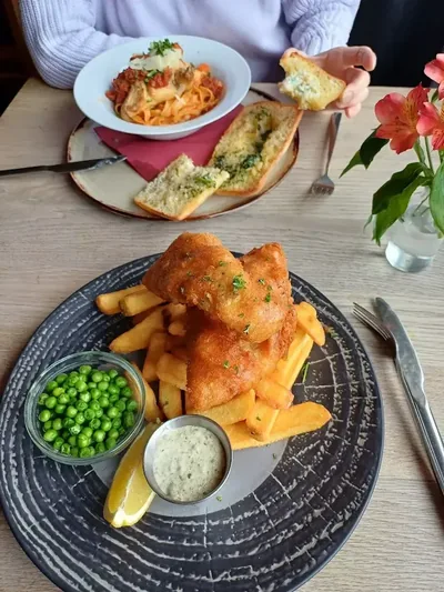 Fish and chips with peas and tartare sauce, and tagliatelle with garlic bread, on a table in the pub